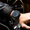 Person wearing a black wristwatch with a blue face in front of a car wheel.